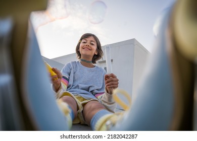 Cute Girl Sitting At The Street Alone And Blowing Soap Bubbles With Wide Smile