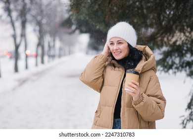 A cute girl smiles and holds a disposable cup of coffee. Adjust the hat in winter under the snow. - Powered by Shutterstock