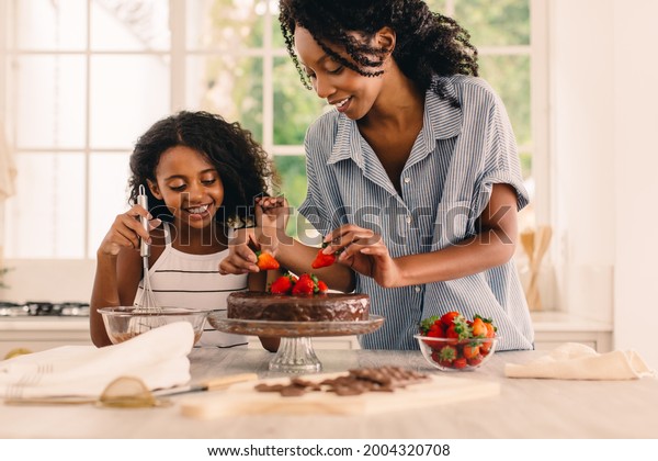 Cute girl standing by her mother in kitchen\
making cake. Mother and happy daughter garnishing chocolate cake\
with fresh strawberries in\
kitchen.