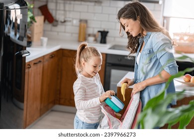 The cute girl is standing next to her caring young mother, who is preparing her to go to school giving her a fruit snack in a lunch box - Powered by Shutterstock