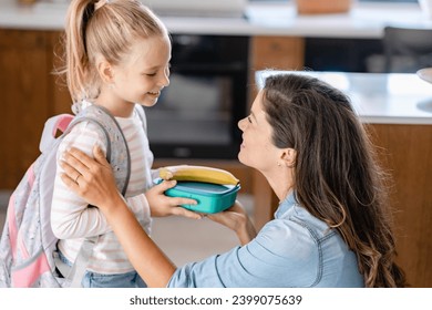 The cute girl is standing next to her caring young mother, who is preparing her to go to school giving her a fruit snack in a lunch box - Powered by Shutterstock