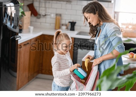 The cute girl is standing next to her caring young mother, who is preparing her to go to school giving her a fruit snack in a lunch box