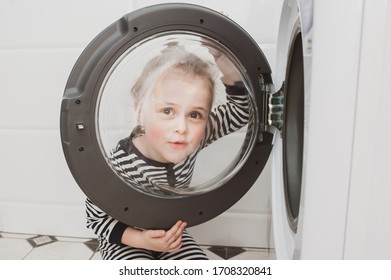 A cute girl in striped clothes peeps out through the round glass door of the washing machine. Cleanliness and Household Concept