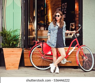 Cute girl in a summer dress, denim jacket, sunglasses stands with red vintage bicycle in a European city. Sunny summer. On background surf cafe.