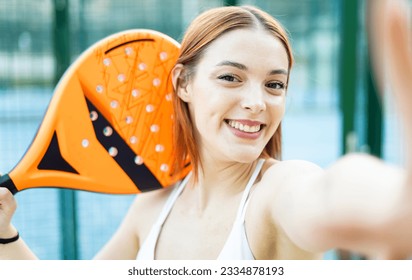 A cute girl is taking a picture with the mobile phone while posing with a paddle racket. Concept of women playing paddle.