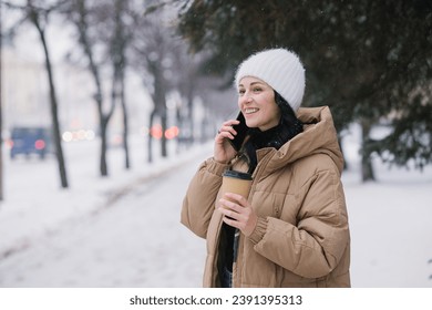 A cute girl is talking on the phone outside in winter and smiling. Hold a disposable cup of coffee - Powered by Shutterstock