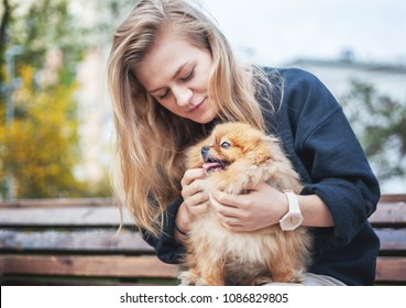 Cute girl teenager with blond hair playing with her puppy Pomeranian Spitz on her hands on a bench in the park