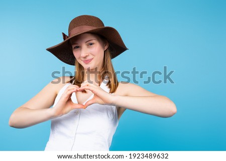 A cute girl tourist isolated on a blue background in a summer hat shows a heart from her hands to the camera. High quality photo