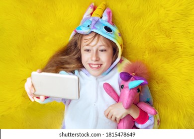 Cute Girl In Unicorn Pajamas With Smartphone In Hand Take Selfies And Having Fun Lying On Floor. Top View Over Yellow Background