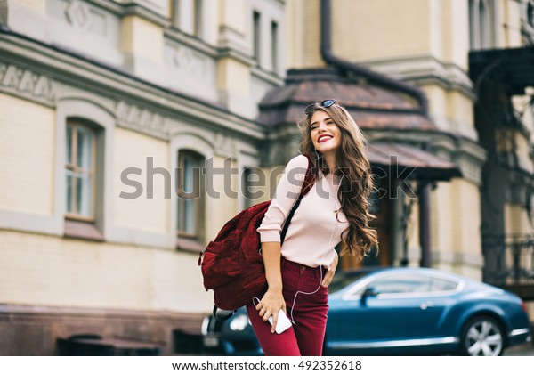 Cute girl with\
vinous bag is walking in the city. She has long hair, listening to\
music and smiling to side.