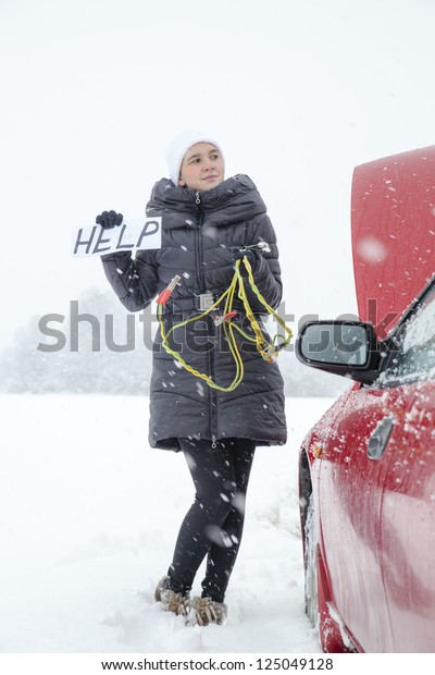Cute girl waiting for help on the road holding HELP\
sign, she needs a battery boost for her car, on very cold, snowy\
winter day