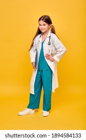 Cute Girl Wearing A Medical Coat And Stethoscope With A Clipboard, Standing On Yellow Background, Full Length. Choosing A Future Profession For A Child