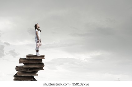 Cute girl wearing pajamas with toy bear in hand standing on pile of books - Shutterstock ID 350132756