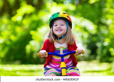 Cute Girl Wearing Safety Helmet Riding Her Tricycle In Sunny Summer Park. Kids Ride Bicycle. First Bike For Little Child. Active Toddler Kid Playing And Cycling Outdoors. Kids Play In The Garden.