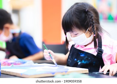 Cute Girl Wearing A White Cloth Face Mask. Students Were In The Water Painting Class. Pupil Kid Wear An Black Apron. Happy Child Learn Arts And Crafts On Canvas. Children Use A Paintbrush To Paint.