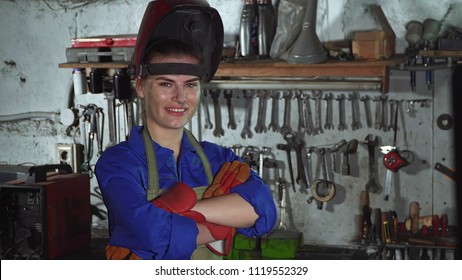 Cute Girl Welder In Uniform In The Garage In The Background Of The Workbench . Women's Work