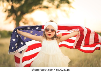 Cute Girl In White Headband And Dress Holds Up American Flag In A Field