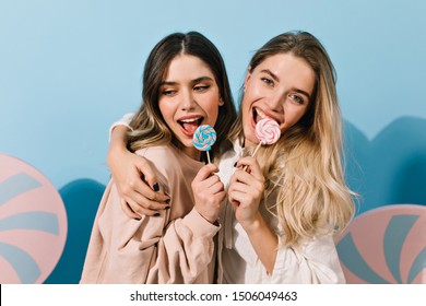 Cute girls with candies embracing on blue background. Studio shot of lovely european women with lollipops.