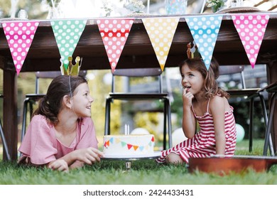 Cute girls with paper crowns eating birthday cake under table. Birthday garden party for children. - Powered by Shutterstock