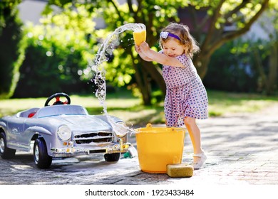 Cute Gorgeous Toddler Girl Washing Big Old Toy Car In Summer Garden, Outdoors. Happy Healthy Little Child Cleaning Car With Soap And Water, Having Fun With Splashing And Playing With Sponge.