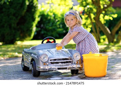 Cute Gorgeous Toddler Girl Washing Big Old Toy Car In Summer Garden, Outdoors. Happy Healthy Little Child Cleaning Car With Soap And Water, Having Fun With Splashing And Playing With Sponge.