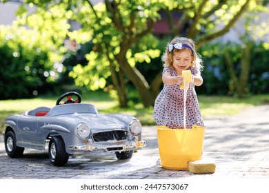 Cute gorgeous toddler girl washing big old toy car in summer garden, outdoors. Happy healthy little child cleaning car with soap and water, having fun with splashing and playing with sponge. - Powered by Shutterstock