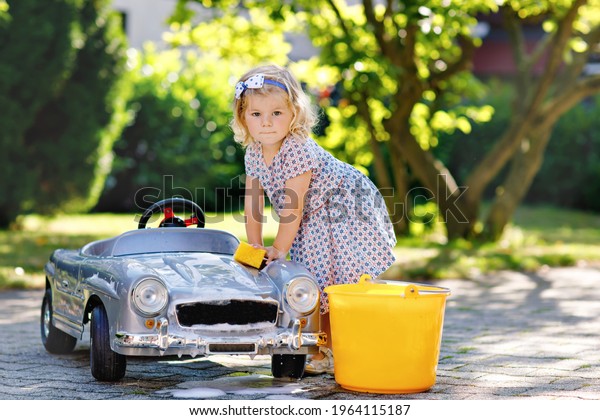 Cute gorgeous toddler girl washing big old toy\
car in summer garden, outdoors. Happy healthy little child cleaning\
car with soap and water, having fun with splashing and playing with\
sponge.