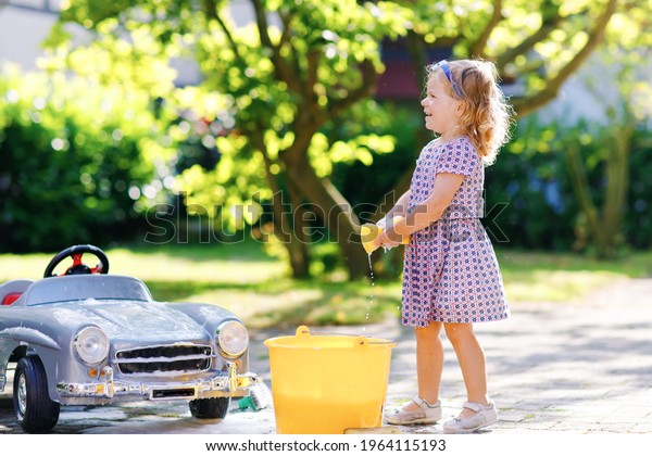 Cute gorgeous toddler girl washing big old toy
car in summer garden, outdoors. Happy healthy little child cleaning
car with soap and water, having fun with splashing and playing with
sponge.