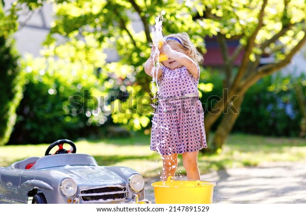 Cute gorgeous toddler girl washing big old toy\
car in summer garden, outdoors. Happy healthy little child cleaning\
car with soap and water, having fun with splashing and playing with\
sponge.