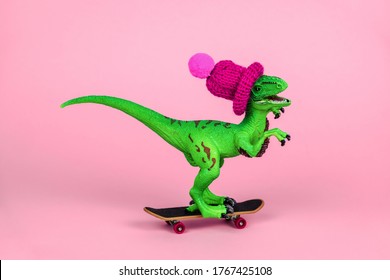 cute green toy dinosaur in knitted hat driving skateboard on a pink background, funny greeting card