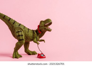 Cute green toy dinosaur speaking on retro phone on pink background. Copy space. 