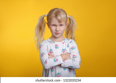 Cute Grumpy Angry Caucasian Blonde Girl With Blue Eyes In White Dress Posing In Studio On Yellow Background With Arms Crossed On Her Chest. Kid Child Expressing Negative Emotion.