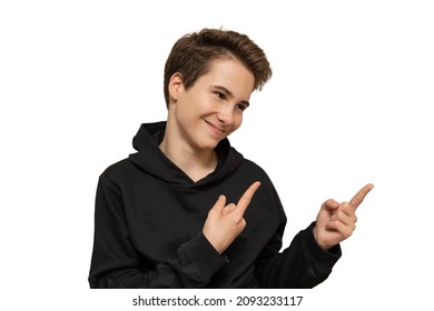 Cute Guy 13-14 Years Old Caucasian In A Black Hoodie Smiles And Shows To Free Space, Close-up Portrait Isolated On White Background For Mockup