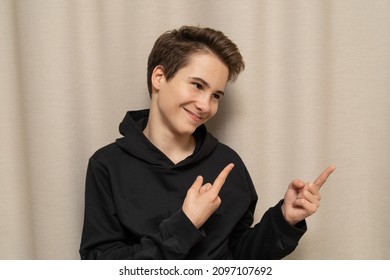 Cute Guy 13-14 Years Old Caucasian In A Black Hoodie Smiles And Shows To Free Space, Close-up Portrait Isolated On Beige Background For Mockup