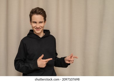 Cute Guy 13-14 Years Old European German In A Black Hoodie Smiles And Shows To Free Space, Close-up Portrait Isolated On Beige Background For Mockup