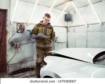 Cute Guy In Black Glasses With Black Gloves Displays The Shape Of The Hood For The Subsequent Painting Of The Car. Guy - A House Painter Working On A Car In The Garage