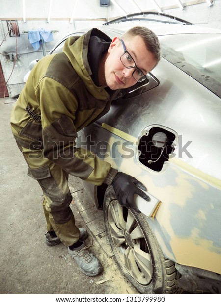 Cute guy in black glasses\
with black gloves displays the shape of the hood for the subsequent\
painting of the car. guy - a house painter working on a car in the\
garage