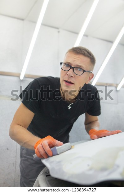 cute guy in black glasses\
with orange gloves displays the shape of the hood for the\
subsequent painting of the car. guy - a house painter working on a\
car in the garage