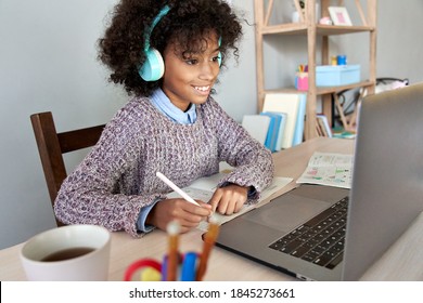 Cute happy african school kid girl wearing headphones virtual distance learning online watching remote digital class lesson looking at laptop computer studying at home sitting at desk, writing notes.
