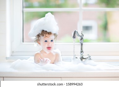 Cute Happy Baby Girl With Big Blue Eyes Playing With Water And Foam In A Kitchen Sink Next To A Window