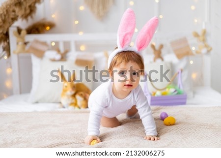 cute happy baby girl in bunny ears on the bed at home with a rabbit and painted eggs, easter concept, funny Easter baby is waiting for the holiday and rejoicing