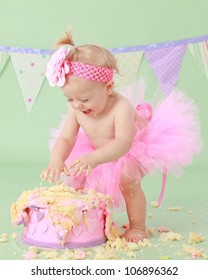 Cute happy blond baby girl in pink tutu and flower head band standing on green background by smashed double tier heart decorated pink fondant iced cake with dirty sticky hands from messy crumb cake