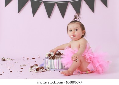 Cute happy brunette baby girl in pink tutu sitting on background by her double tier pink and purple fondant iced birthday party cake reaching out touching and destroying it looking at camera surprised