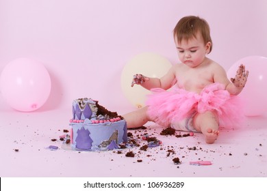 Cute happy brunette baby girl in pink tutu sitting on pink background by her birthday double tier pink and purple butter iced chocolate cake with dirty sticky hands looking at her destroyed mess