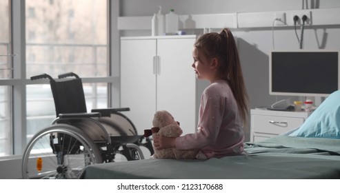 Cute Happy Child Alone In Hospital Room Sitting On Bed With Teddy Bear. Side View Of Disabled Little Girl Sitting On Bed In Hospital Ward Waiting For Medical Assistance