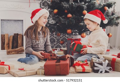 Cute Happy Excited Children, Boy And Girl In Santa Hats Unwrap Christmas Present Box In Beautiful Room. Sister And Brother Open Xmas Gifts Near Decorated Tree And Fireplace. Family Holiday