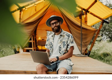 Cute happy freelancer guy using laptop on a sunny day away from the city on a campsite with free wifi. Lifestyle concept. Stay connected. Remote work in nature