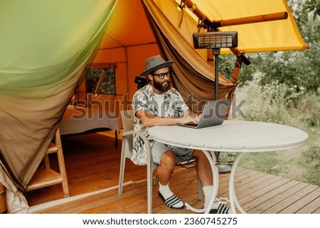 Cute happy freelancer guy using laptop on a sunny day away from the city on a campsite with free wifi. Lifestyle concept. Stay connected. Remote work in nature