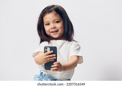 A Cute, Happy Girl Of Preschool Age Stands On A Light Background In A White T-shirt With A Fashionable Smartphone And Looks Into It With Enthusiasm