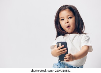 A Cute, Happy Girl Of Preschool Age Stands On A Light Background In A White T-shirt With A Fashionable Smartphone And Looks Into It With Enthusiasm